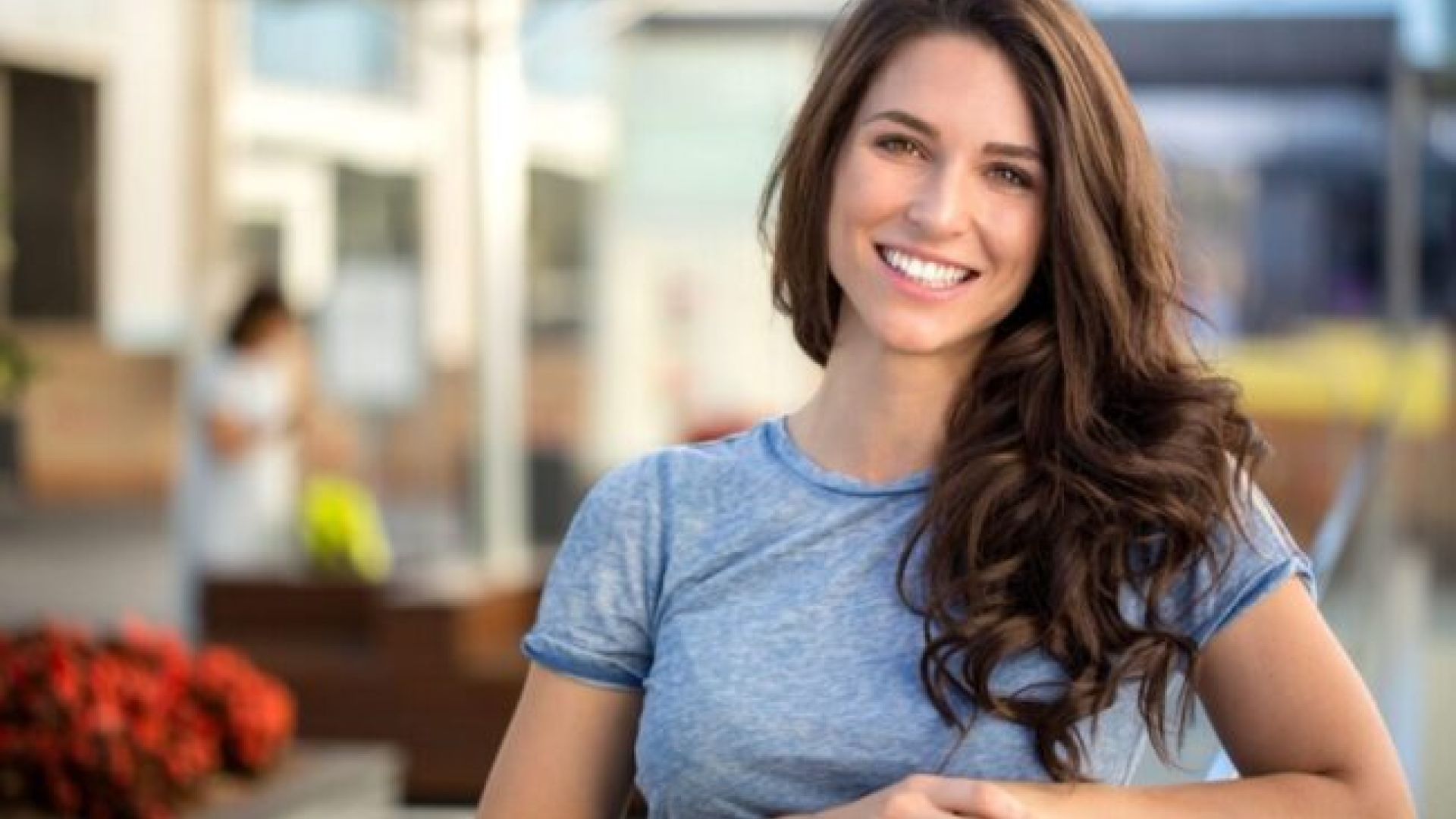 Smiling woman with long brown hair wearing a blue shirt, sitting outdoors in a sunny setting.
