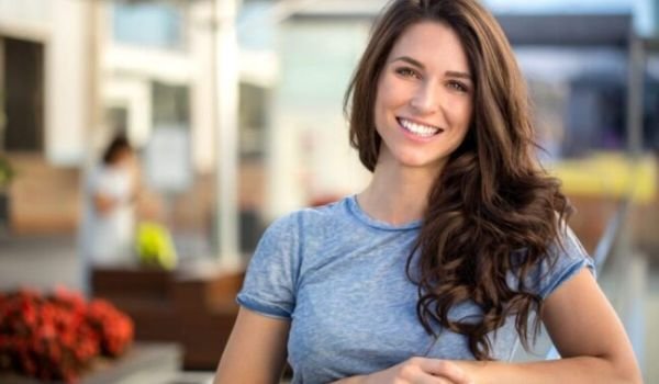 Smiling woman with long brown hair wearing a blue shirt, sitting outdoors in a sunny setting.