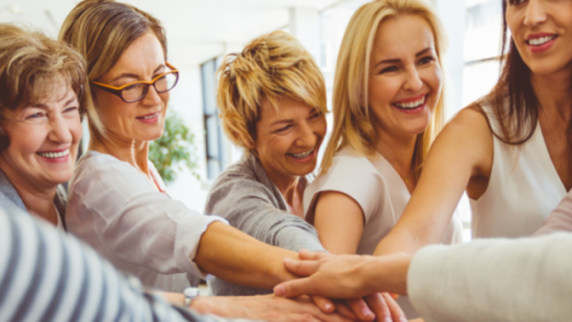 A group of smiling women standing together with their hands stacked in unity, symbolizing support, strength, and community.