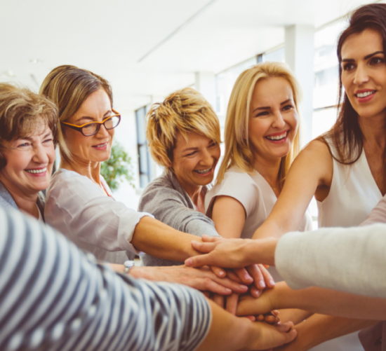 A group of smiling women standing together with their hands stacked in unity, symbolizing support, strength, and community.