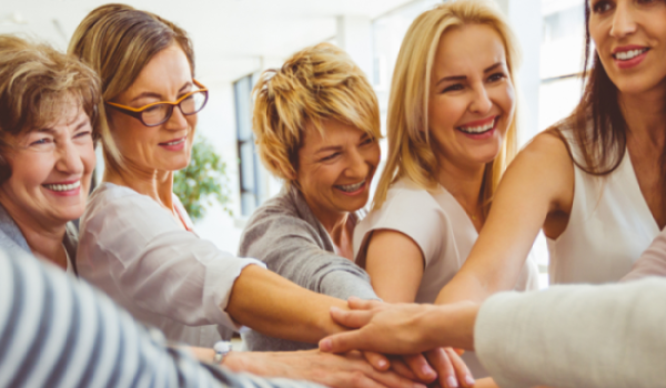 A group of smiling women standing together with their hands stacked in unity, symbolizing support, strength, and community.